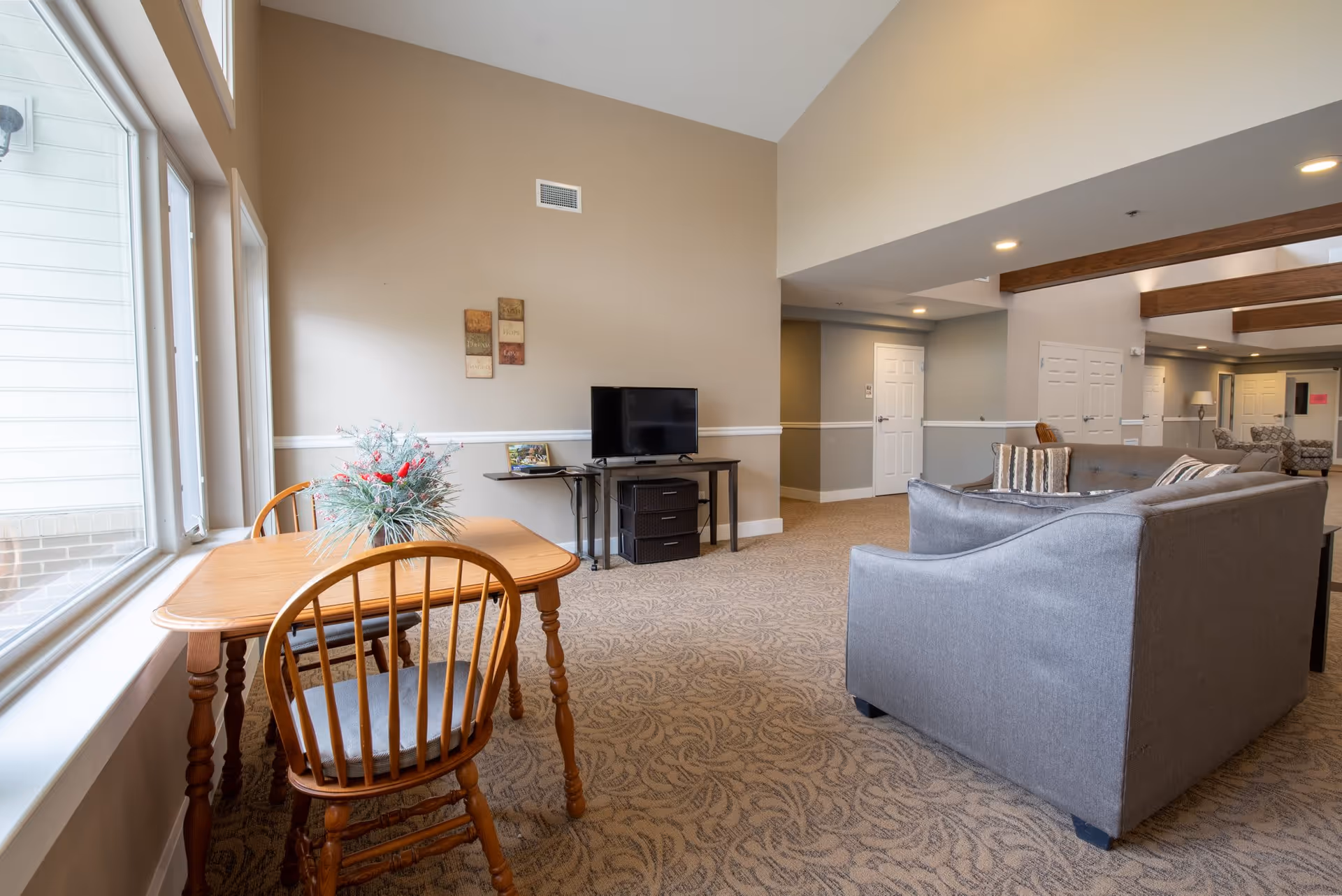 A cozy living room interior featuring a wooden dining table with a floral arrangement, a television on a stand, and comfortable seating. The room has large windows allowing natural light and a neutral color scheme with decorative wall art.