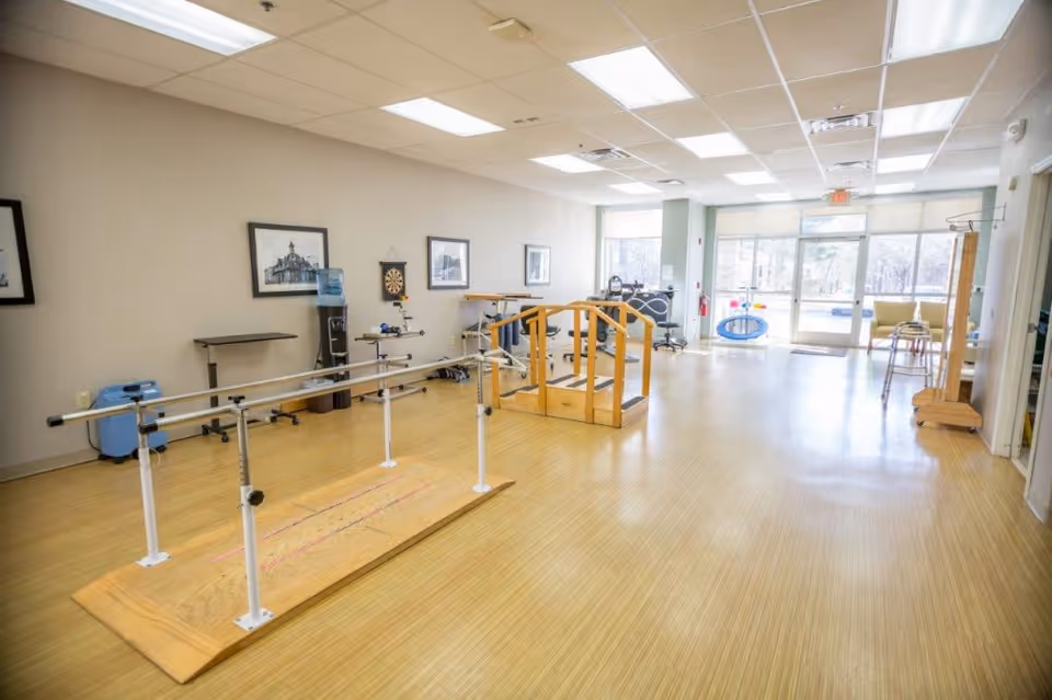 A spacious rehabilitation or physical therapy room with wooden flooring, parallel bars for walking exercises, a small wooden staircase with handrails, exercise equipment, framed pictures on the wall, and large windows letting in natural light.