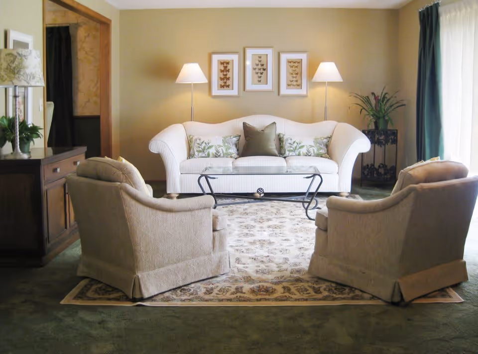 Sunlit living room featuring a white upholstered sofa, two beige armchairs, a glass coffee table, an area rug, and floor lamps.
