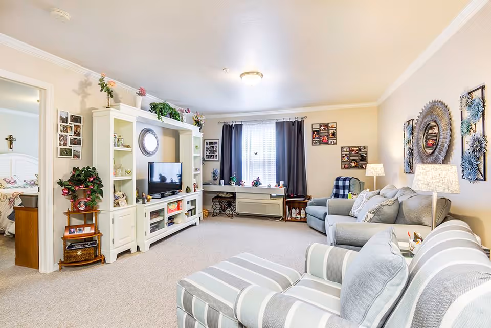 A bright and cozy living room in a retirement community featuring a striped armchair and matching ottoman in the foreground, a gray sofa and armchair with decorative pillows, a white entertainment center with a TV and various decorations, and a window with dark curtains letting in natural light. The walls are adorned with framed photos and decorative wall art. A doorway on the left leads to a bedroom with a white bed and a cross on the wall.