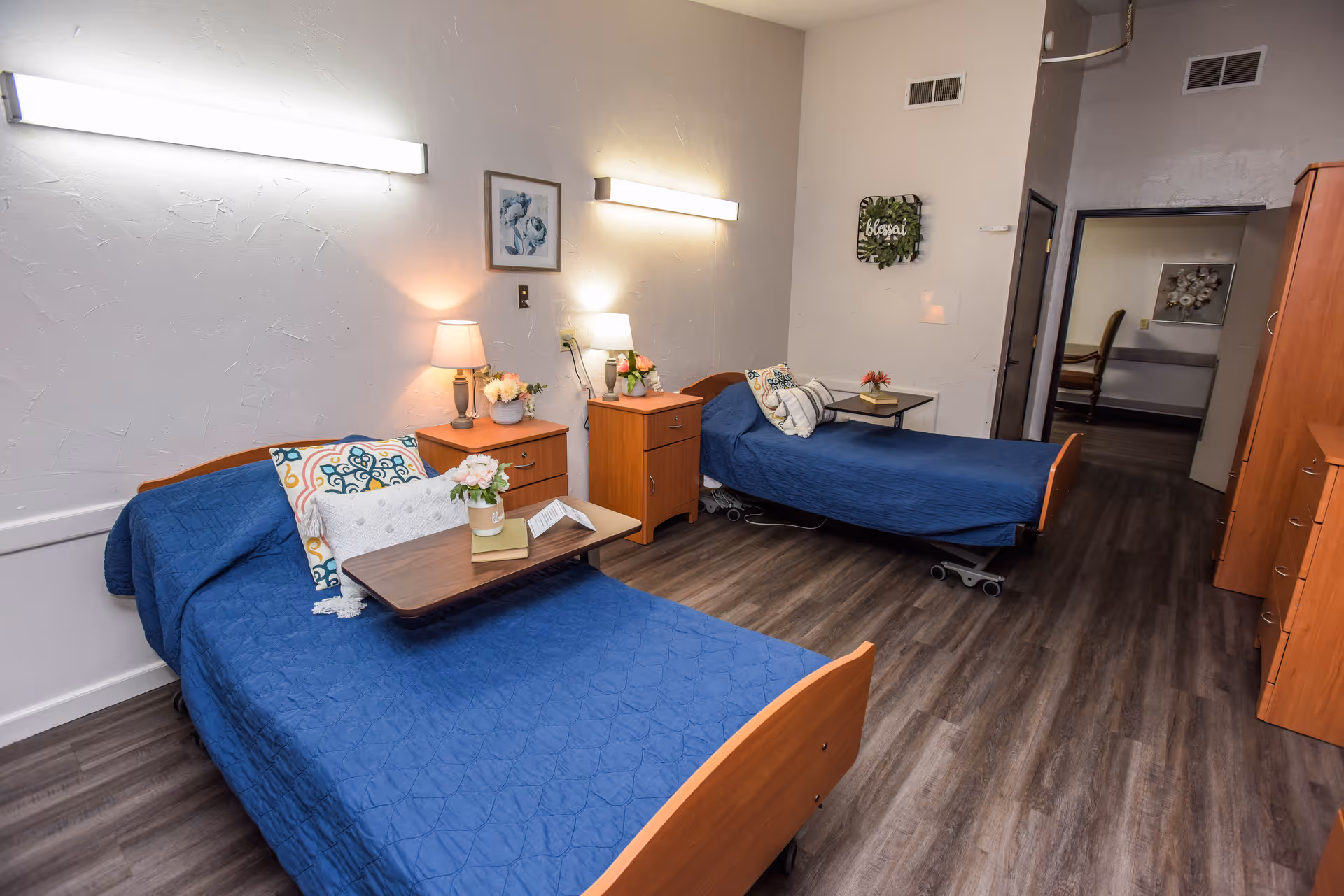 Two single hospital-style beds with blue bedspreads face wooden nightstands and lamps in a shared senior care bedroom.