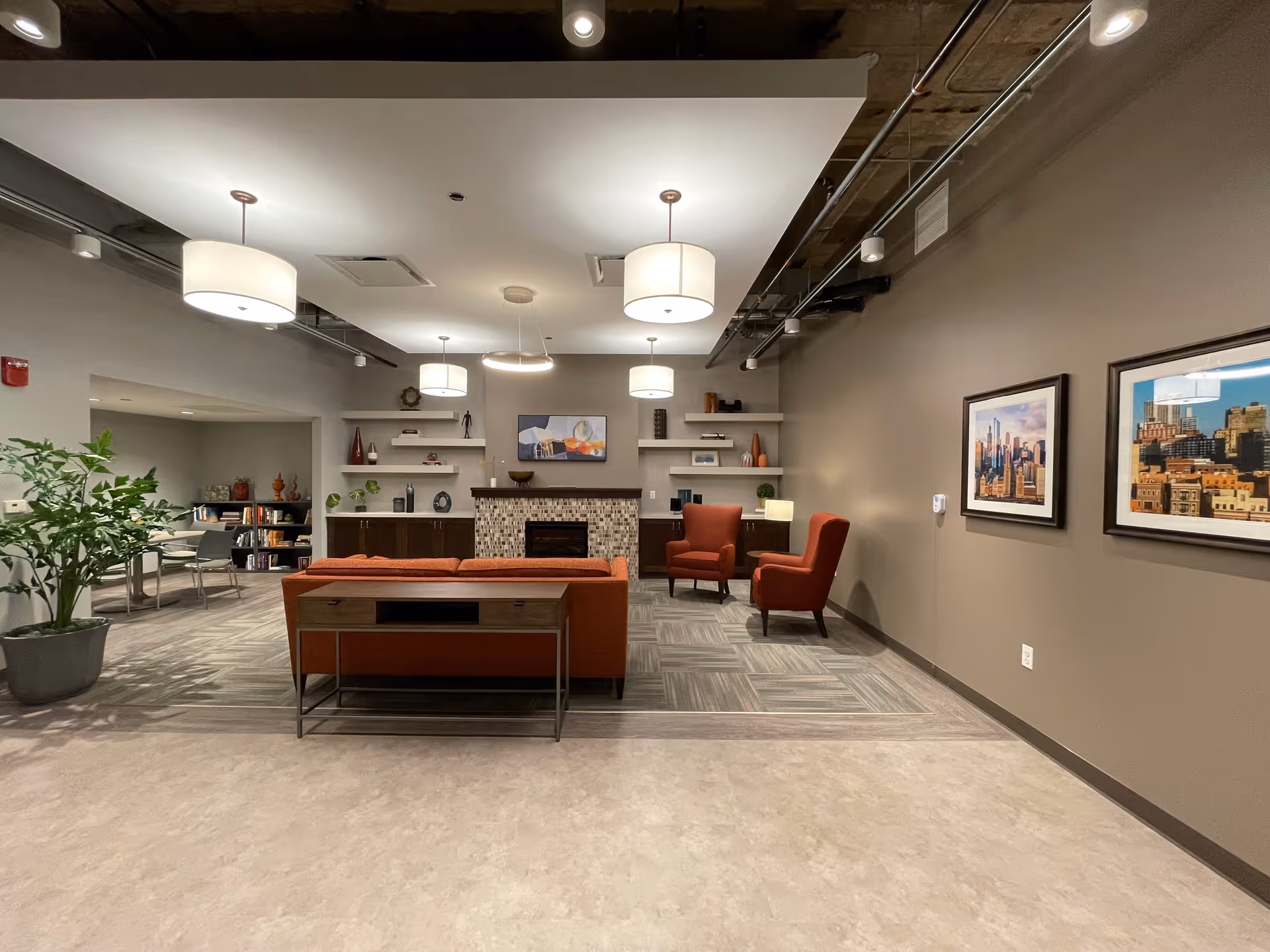 A modern and cozy living room area in Ravenswood Supportive Living featuring an orange sofa, two orange armchairs, a fireplace with a mosaic tile surround, built-in shelves with decorative items, and framed cityscape artwork on the wall. The room has a mix of carpet and tile flooring, ceiling lights, and an adjacent area with a table and chairs.