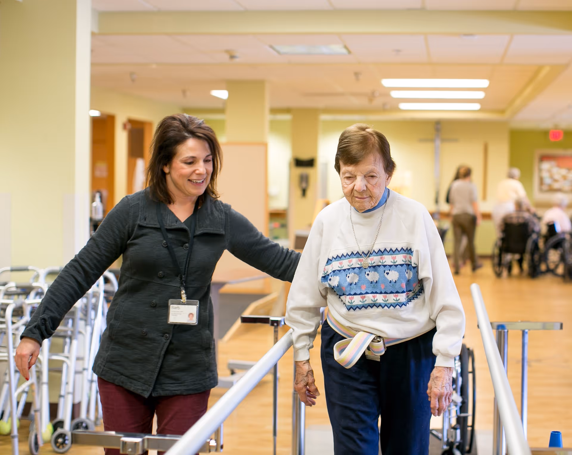 A female caregiver assists an elderly woman as she walks between parallel bars in a rehabilitation or therapy area of a senior living facility. In the background, other elderly individuals and caregivers are visible in a spacious, well-lit room with wooden floors.