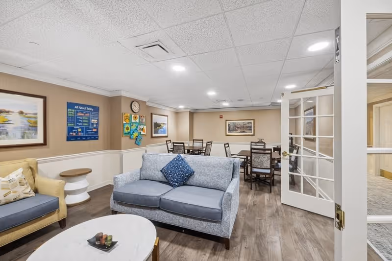 A cozy common area in a senior living facility featuring a light blue sofa with a blue patterned pillow, a yellow armchair with a patterned pillow, a round white coffee table with decorative items, and a dining area with multiple tables and chairs in the background. The walls are decorated with framed artwork and a bulletin board, and the room has wood flooring and a white ceiling with recessed lighting.