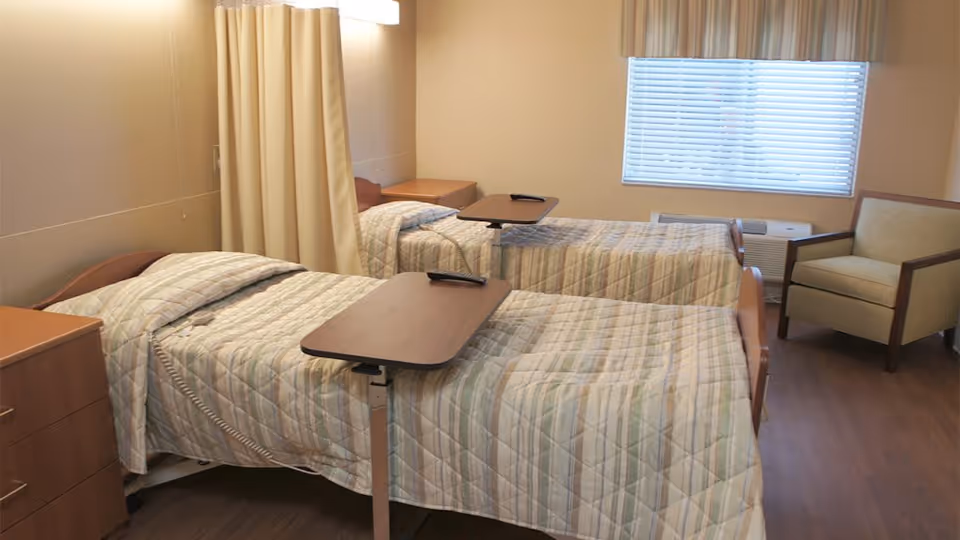 A nursing facility bedroom with two hospital-style beds, overbed tables, a privacy curtain, a dresser and a chair by a window.