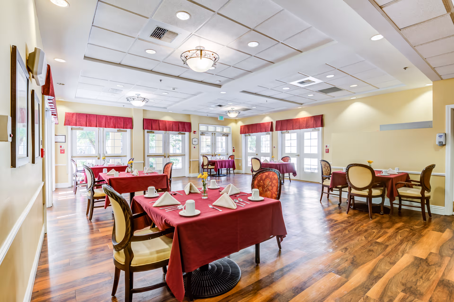 A bright dining room in a senior living facility with several tables covered in red tablecloths, each set with white napkins, cups, and silverware. The room has wooden flooring, cream-colored walls, large windows with red valances, and elegant chairs with cushioned seats and backs. Ceiling lights provide additional illumination.