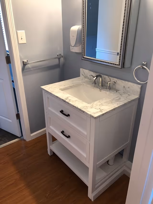Small bathroom with a white vanity topped by a marble countertop and undermount sink, mirrored medicine cabinet, and towel racks against light blue-gray walls.