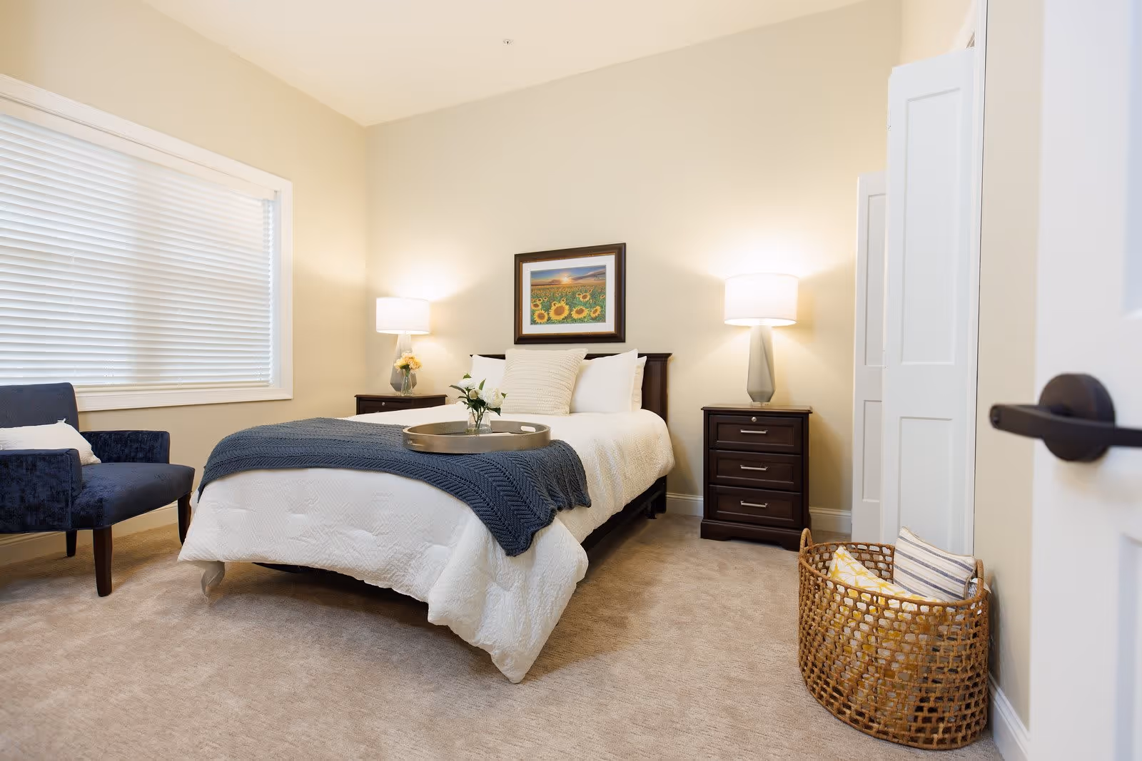 Bright, neatly staged bedroom with a bed, two nightstands and lamps, a blue chair by the window, and a woven basket.