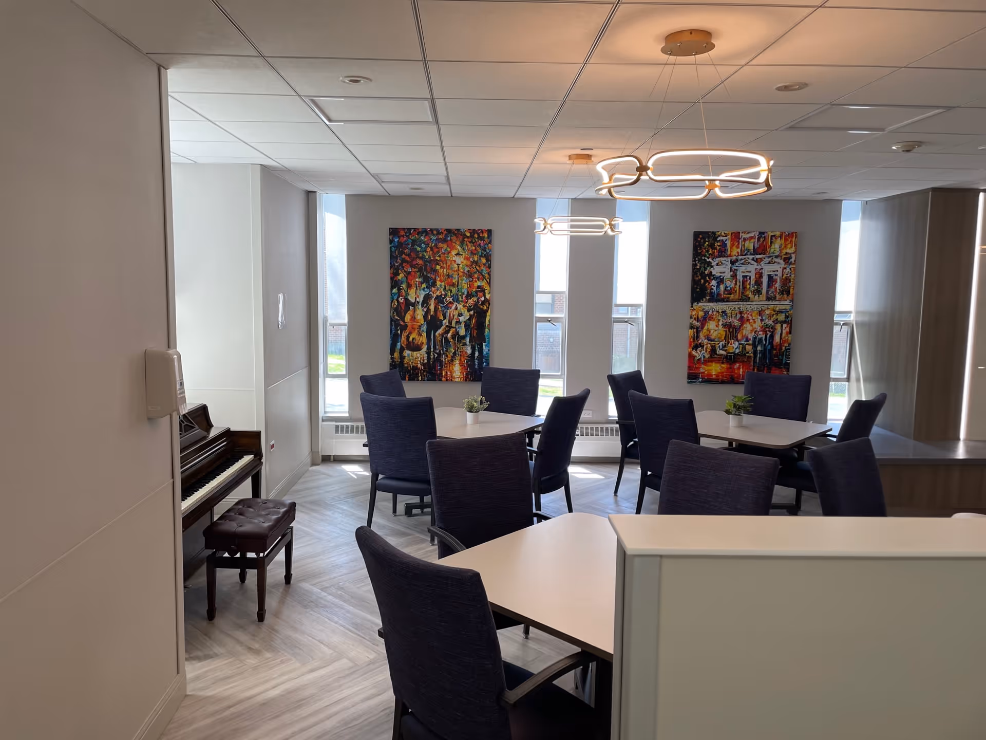 A bright, modern common area with several tables and purple upholstered chairs arranged around them. Two colorful paintings hang on the wall between tall windows letting in natural light. A piano with a matching bench is positioned against the left wall. The ceiling has contemporary circular light fixtures.