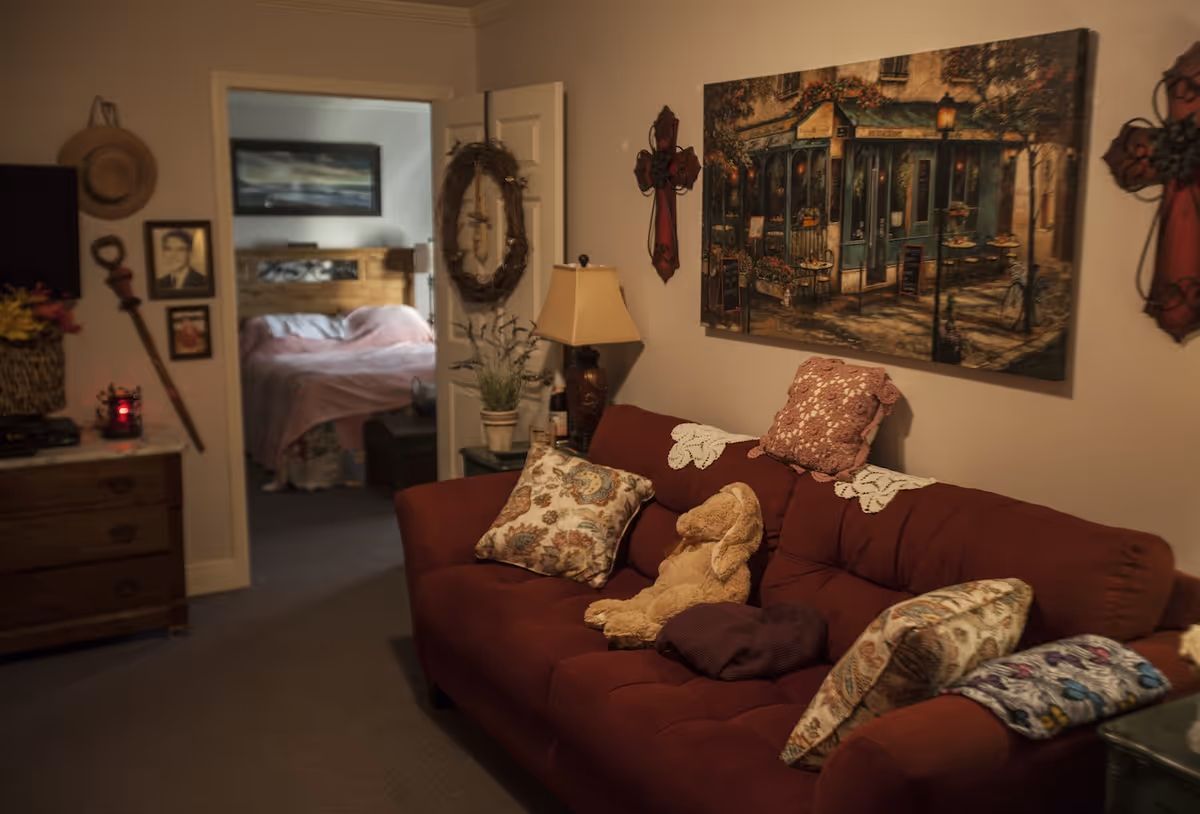 Cozy living room with a red sofa topped with decorative pillows and a stuffed toy, wall art and a lamp, and a doorway leading to a bedroom.
