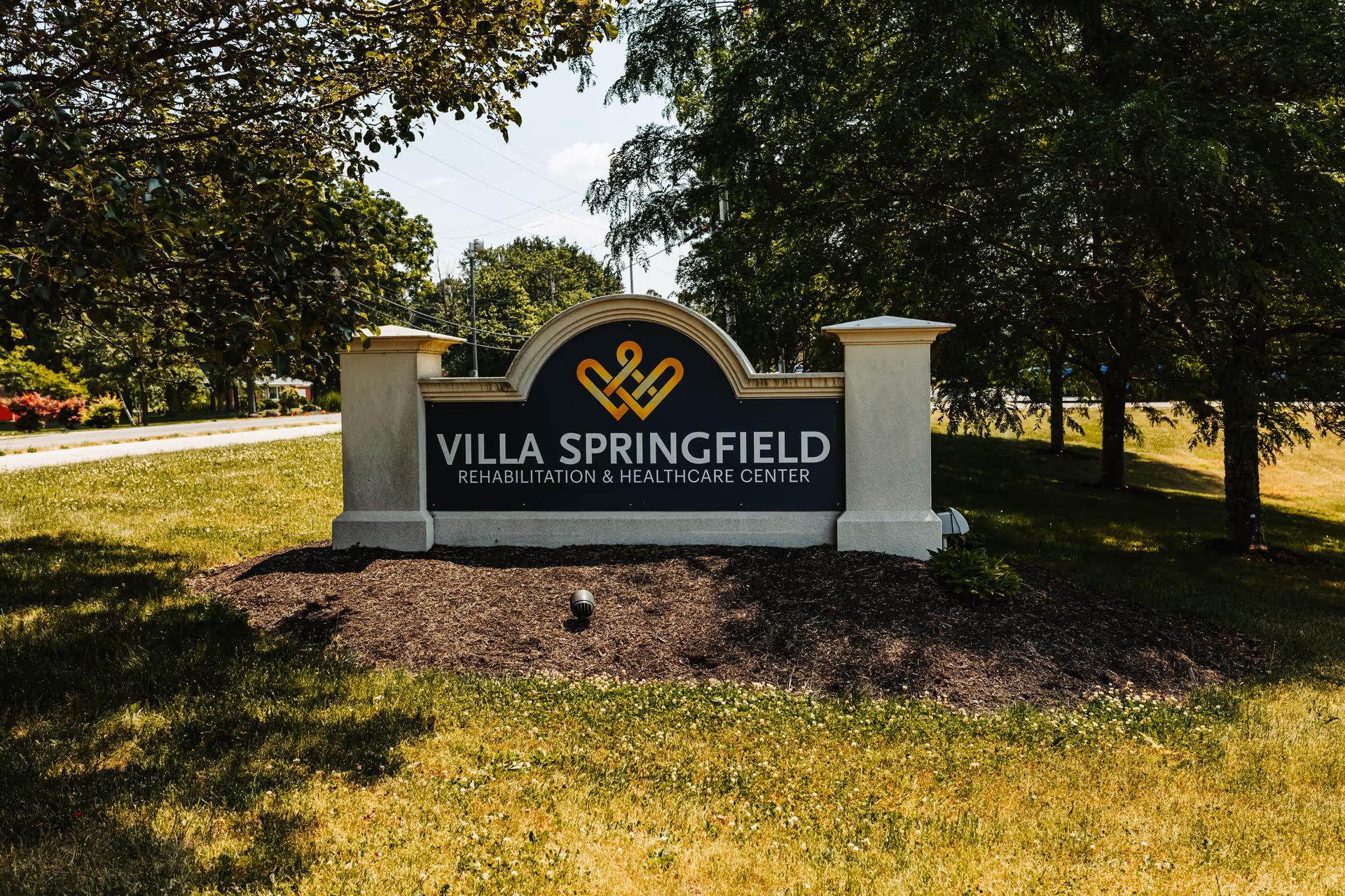 Outdoor view of a sign for Villa Springfield Rehabilitation & Healthcare Center surrounded by grass, trees, and landscaping under a partly cloudy sky.