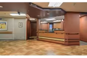 Interior view of a reception or front desk area in a facility with a wooden counter, beige tiled floor, and brown walls. There is a sign above the desk that reads 'Bistro' and a hallway leading to other rooms. The lighting is fluorescent and the space appears clean and organized.