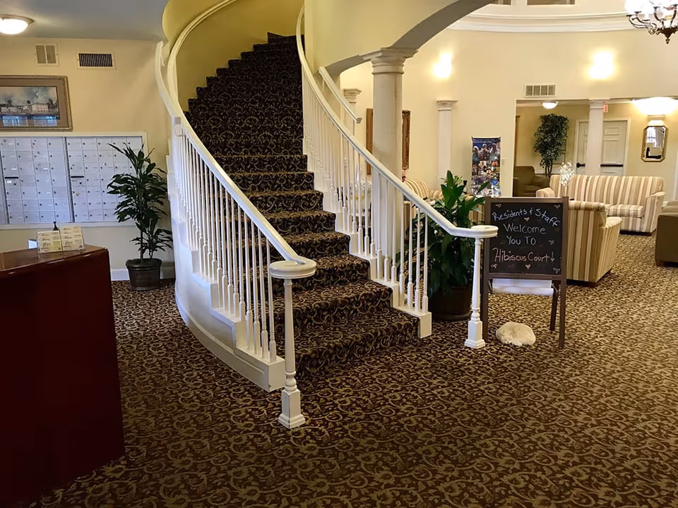 Interior view of a senior living facility lobby with a carpeted curved staircase with white railings in the center. To the right, there is a chalkboard sign welcoming residents and staff to Hibiscus Court, surrounded by potted plants and striped sofas. On the left, there is a reception desk and a wall of mailboxes. The area is well-lit with ceiling lights and decorated with framed artwork and columns.
