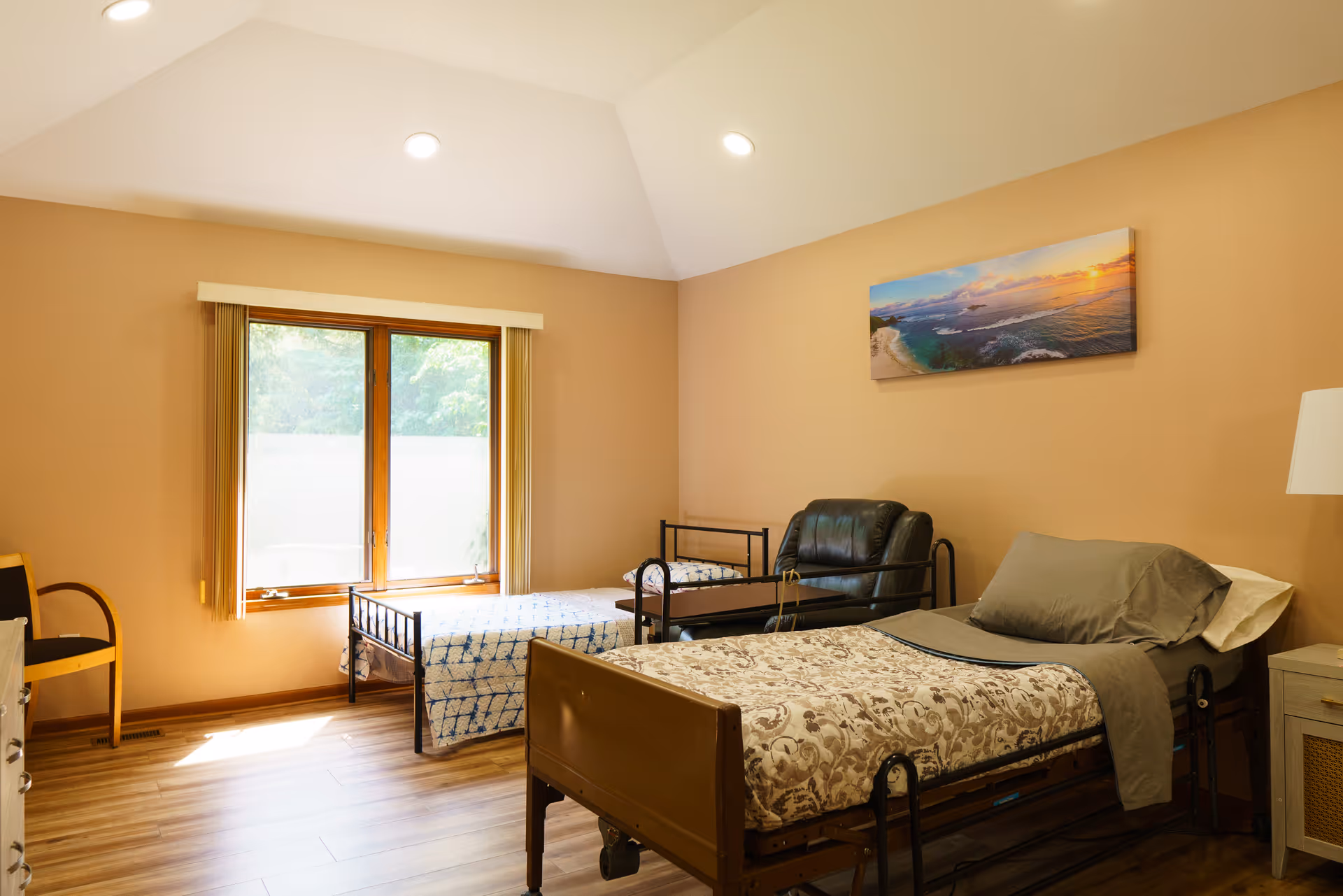 A cozy bedroom in a senior living facility featuring two beds, a chair, and a window with a view.