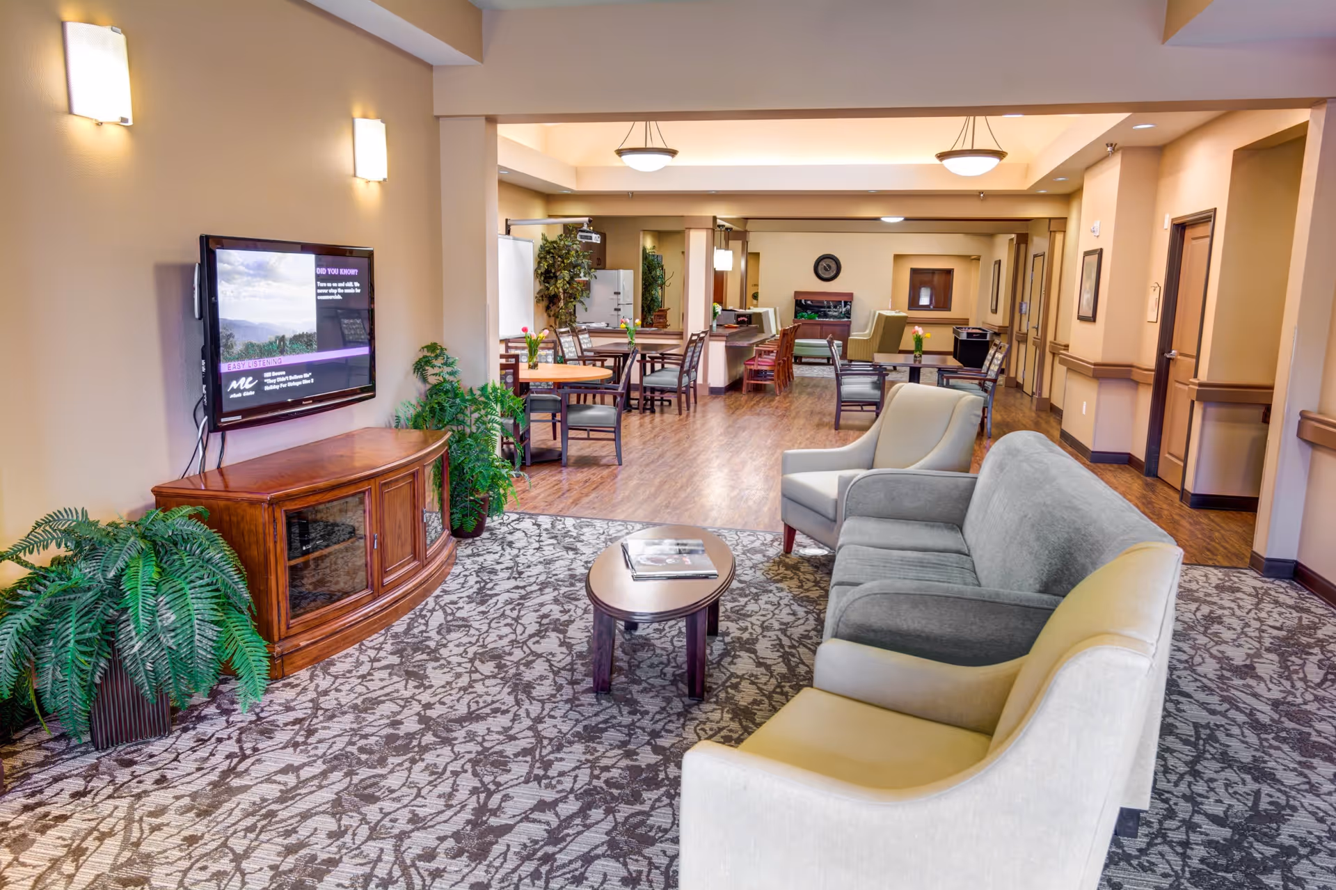 A spacious and well-lit common area in a senior living facility featuring a seating arrangement with a gray sofa, two beige armchairs, a small oval coffee table, and a flat-screen TV mounted on the wall above a wooden cabinet. The room has a patterned carpet, wooden flooring, several tables and chairs in the background, potted plants, and soft wall lighting.