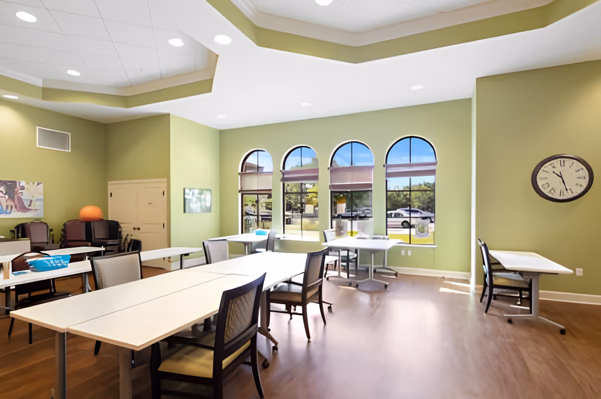 A bright room with light green walls and large arched windows letting in natural light. The room contains several tables and chairs arranged for group seating, with a clock on the wall showing the time as 11:05. The floor is wooden, and there are some stacked chairs and a colorful painting on one wall.