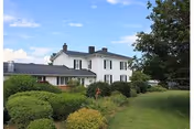 A large white two-story house with a black roof surrounded by green bushes and trees under a blue sky with some clouds.