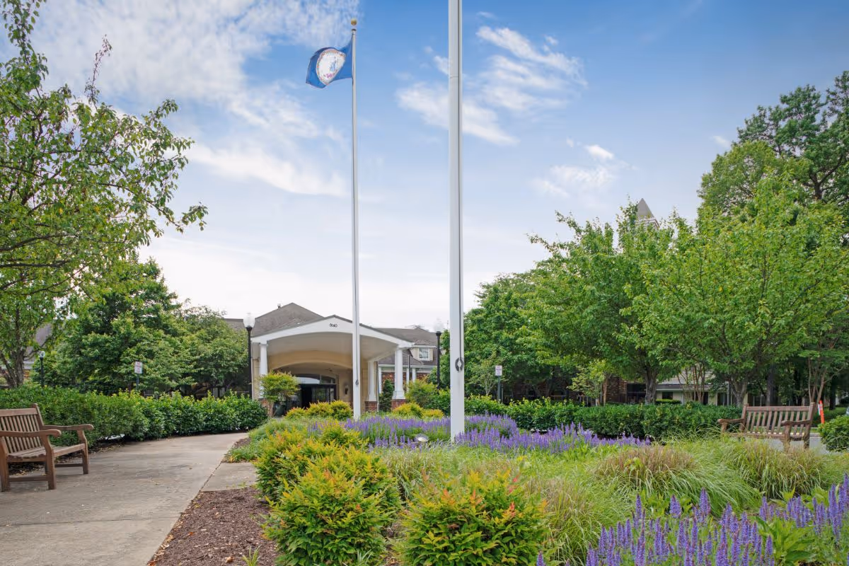 Outdoor view of The Fairfax at Belvoir Woods facility entrance with a pathway lined by green bushes, purple flowers, and trees. Two flagpoles stand in the center of the landscaped garden area, and wooden benches are placed along the pathway.
