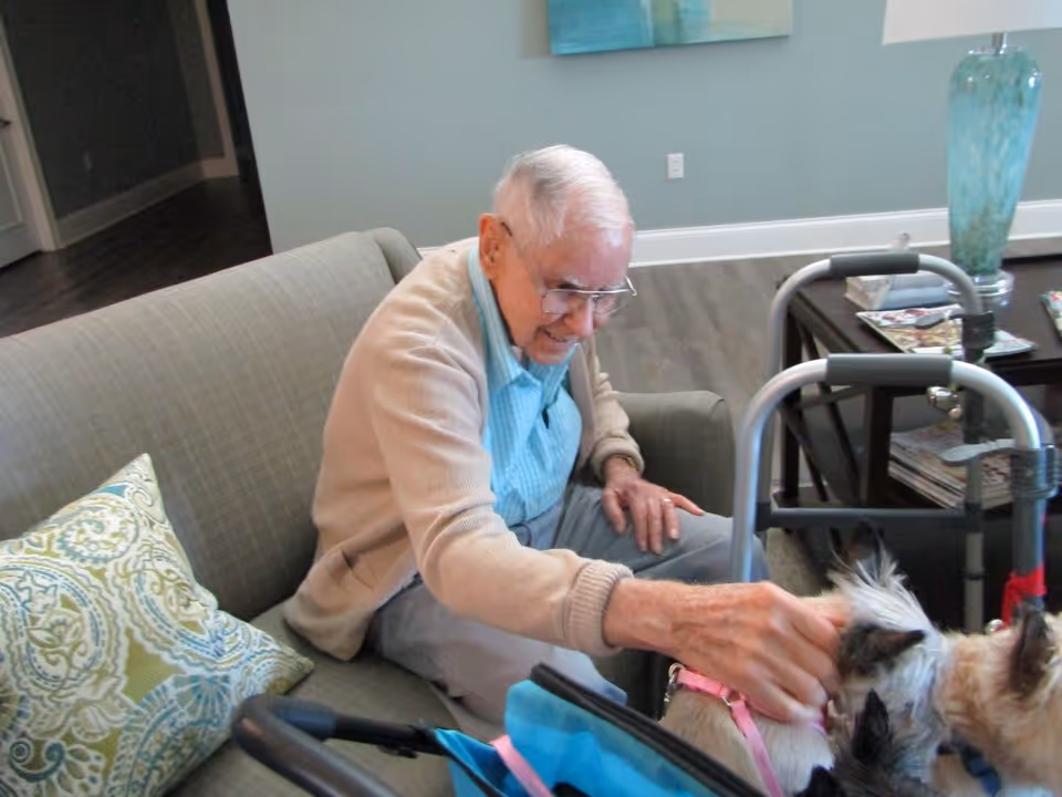 An elderly man sitting on a couch in a living room, smiling and petting two small dogs. A walker is positioned nearby, and a coffee table with magazines and a blue vase is in the background.