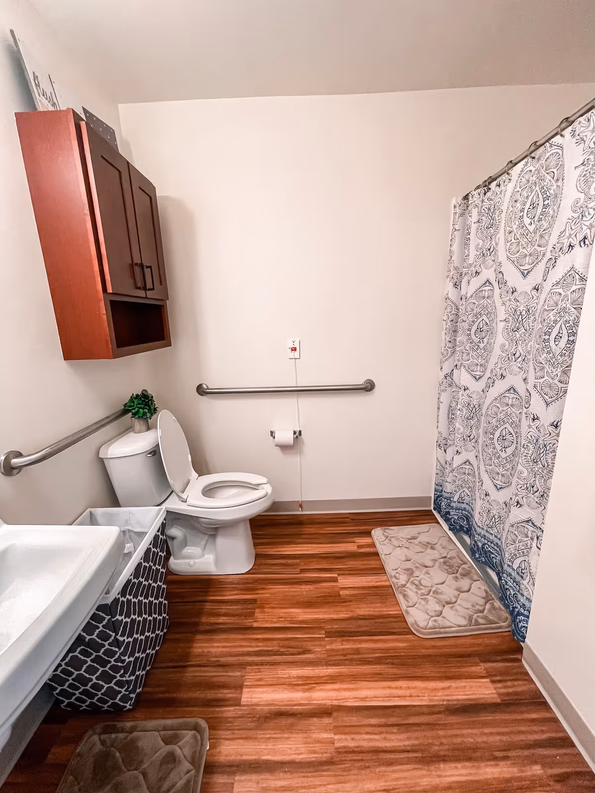A clean bathroom with wooden floor, a white toilet with the lid open, a white sink, a patterned shower curtain, two beige bath mats, a wooden cabinet mounted on the wall, and grab bars on the walls for accessibility.