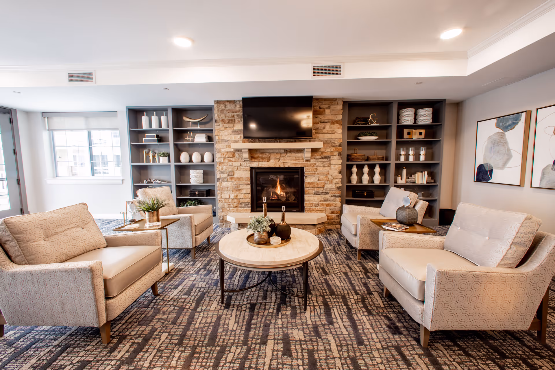 A cozy senior living room with four beige armchairs arranged around a round coffee table. The room features a stone fireplace with a mounted TV above it, built-in shelves on either side filled with decorative items, and two abstract paintings on the wall. The space is well-lit with natural light coming from a window on the left.