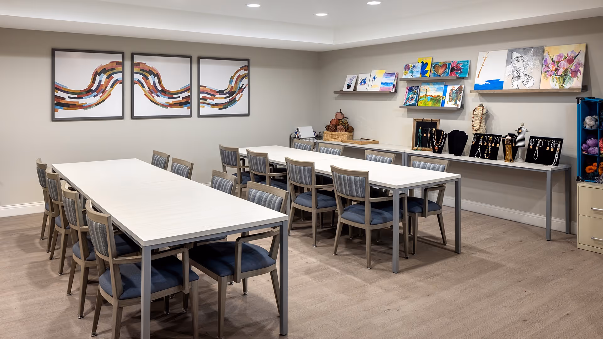 A bright room with two long white tables surrounded by wooden chairs with blue cushions. The walls are decorated with colorful abstract paintings and several small artworks displayed on shelves. There are also jewelry displays and craft supplies on a long table against the wall.