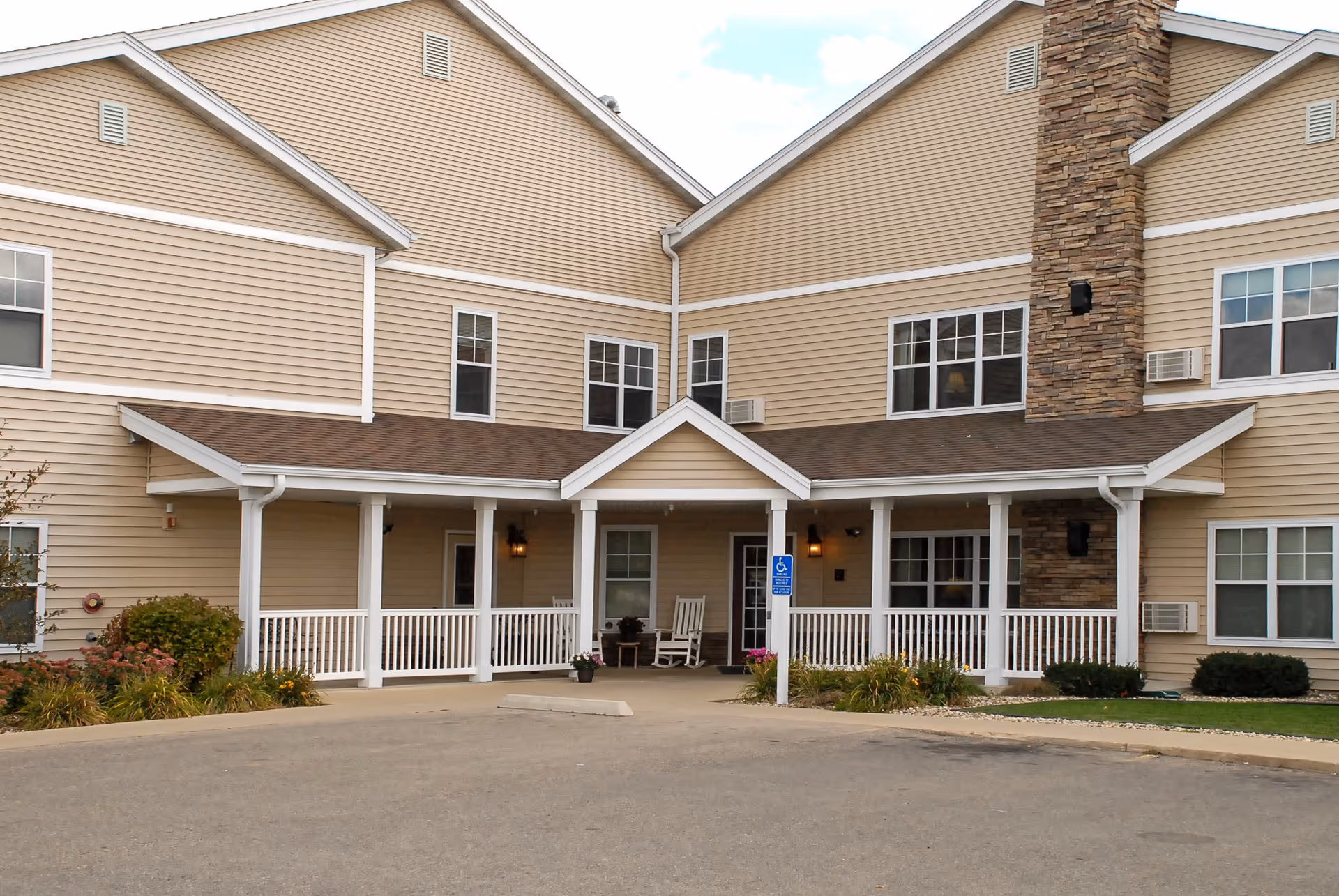 Front entrance of a beige two-story senior living building with a covered porch, white railing, and a driveway.