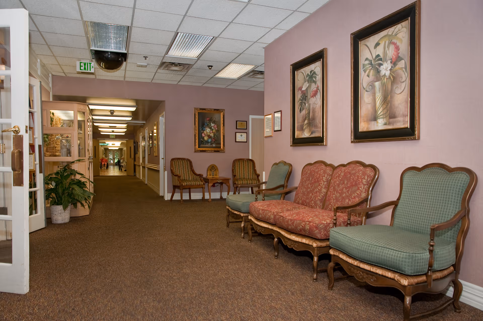 Carpeted hallway in a care center with upholstered chairs along a mauve wall and framed artwork, leading to a distant corridor.