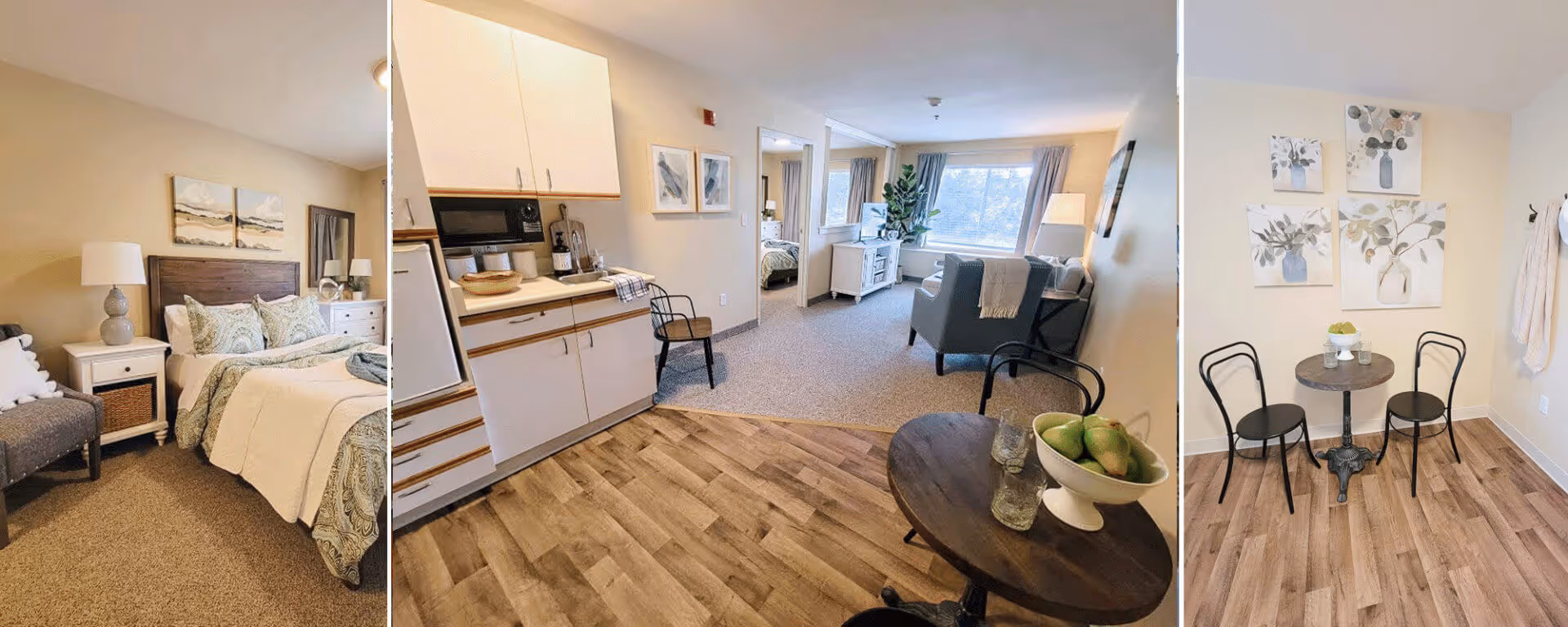 A senior living apartment interior showing a cozy bedroom with a bed, nightstand, and lamp on the left; a small kitchenette with white cabinets and a microwave in the center; a living area with armchairs, a TV, and a large window in the background; and a small dining nook with a round table and two chairs on the right. The space features wood and carpet flooring, neutral walls, and decorative wall art.