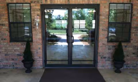 Brick building entrance with double glass doors and two potted shrubs.
