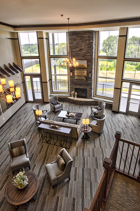A spacious senior living facility common area with large floor-to-ceiling windows letting in natural light. The room features a stone fireplace, several comfortable armchairs and sofas arranged around coffee tables, and modern lighting fixtures. The flooring is carpeted with a patterned design, and there is a wooden staircase visible in the foreground.