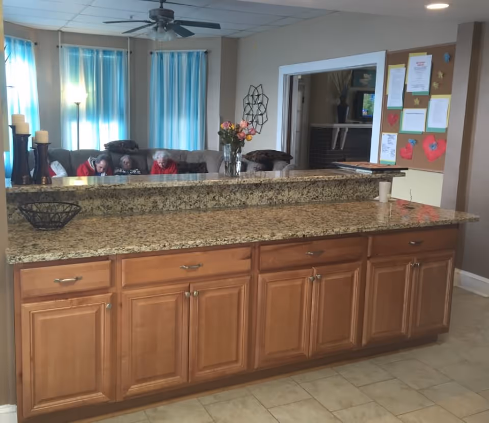View of a granite countertop kitchen island with wooden cabinets in an assisted living facility. In the background, there is a living room area with a sofa where three elderly people are seated. The room has blue curtains, a ceiling fan, a floor lamp, and a bulletin board with various papers and decorations on the wall.