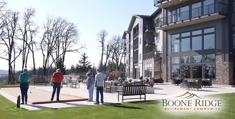 Four people playing bocce ball on a court outside a multi-story retirement community building with large windows and balconies. The area is surrounded by grass and trees under a clear sky.