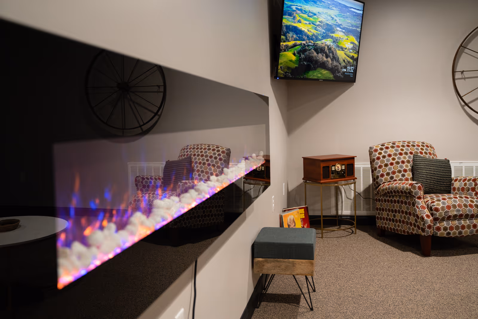 Cozy lounge with patterned armchairs, an electric fireplace, a wall-mounted TV, and a small side table with decor.
