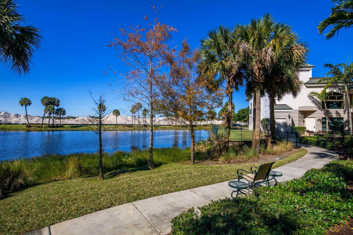 Lakeside walking path with benches, palm trees, and a senior living building beside a pond under a clear blue sky.