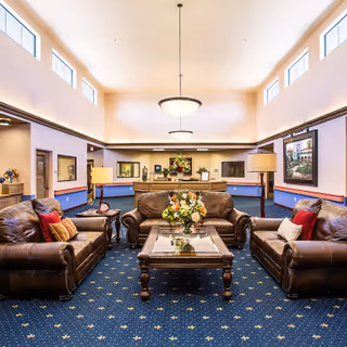 A spacious and well-lit common area with three brown leather sofas arranged around a wooden coffee table with a floral centerpiece. The room has blue carpet with a star pattern, high ceilings with multiple windows near the ceiling, and two standing lamps. There are framed pictures on the walls and a reception desk in the background.