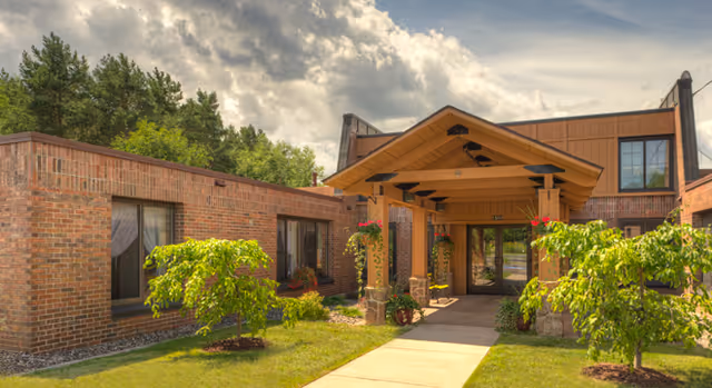 Exterior view of a brick building with a covered entrance supported by stone pillars, surrounded by green shrubs and small trees under a partly cloudy sky.