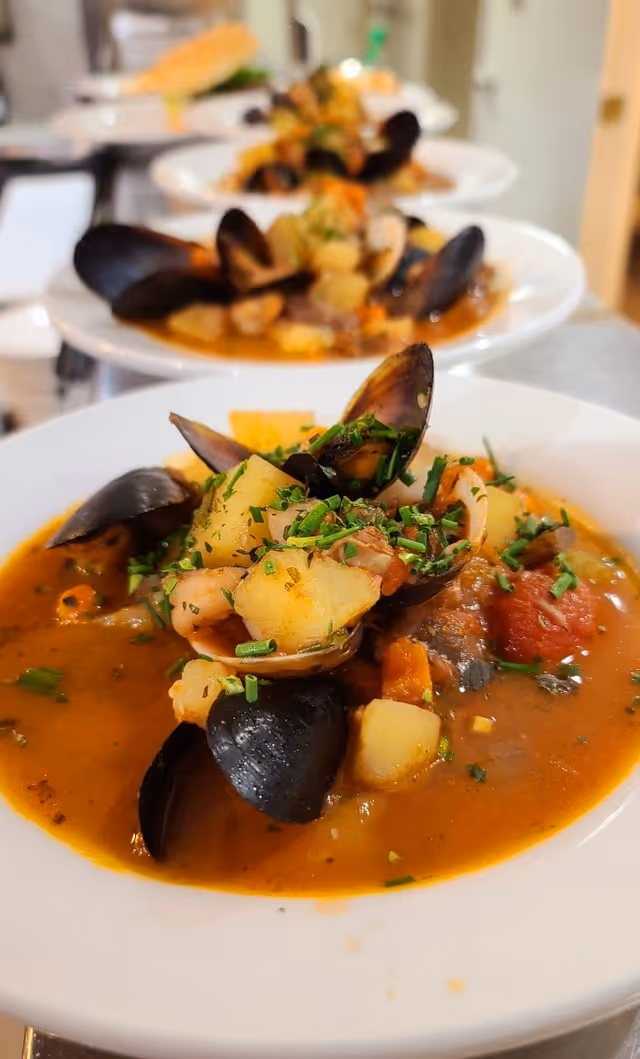 Close-up view of a bowl of seafood stew with mussels, potatoes, and herbs, with several similar bowls lined up in the background.