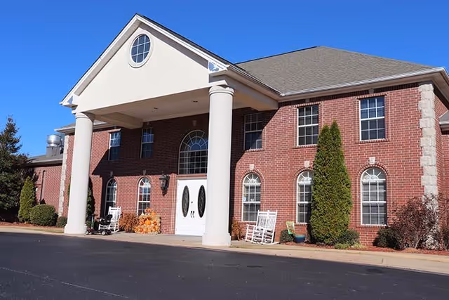 Exterior view of a two-story brick building with white columns at the entrance, white double doors, multiple windows, and some landscaping including bushes and small trees under a clear blue sky.