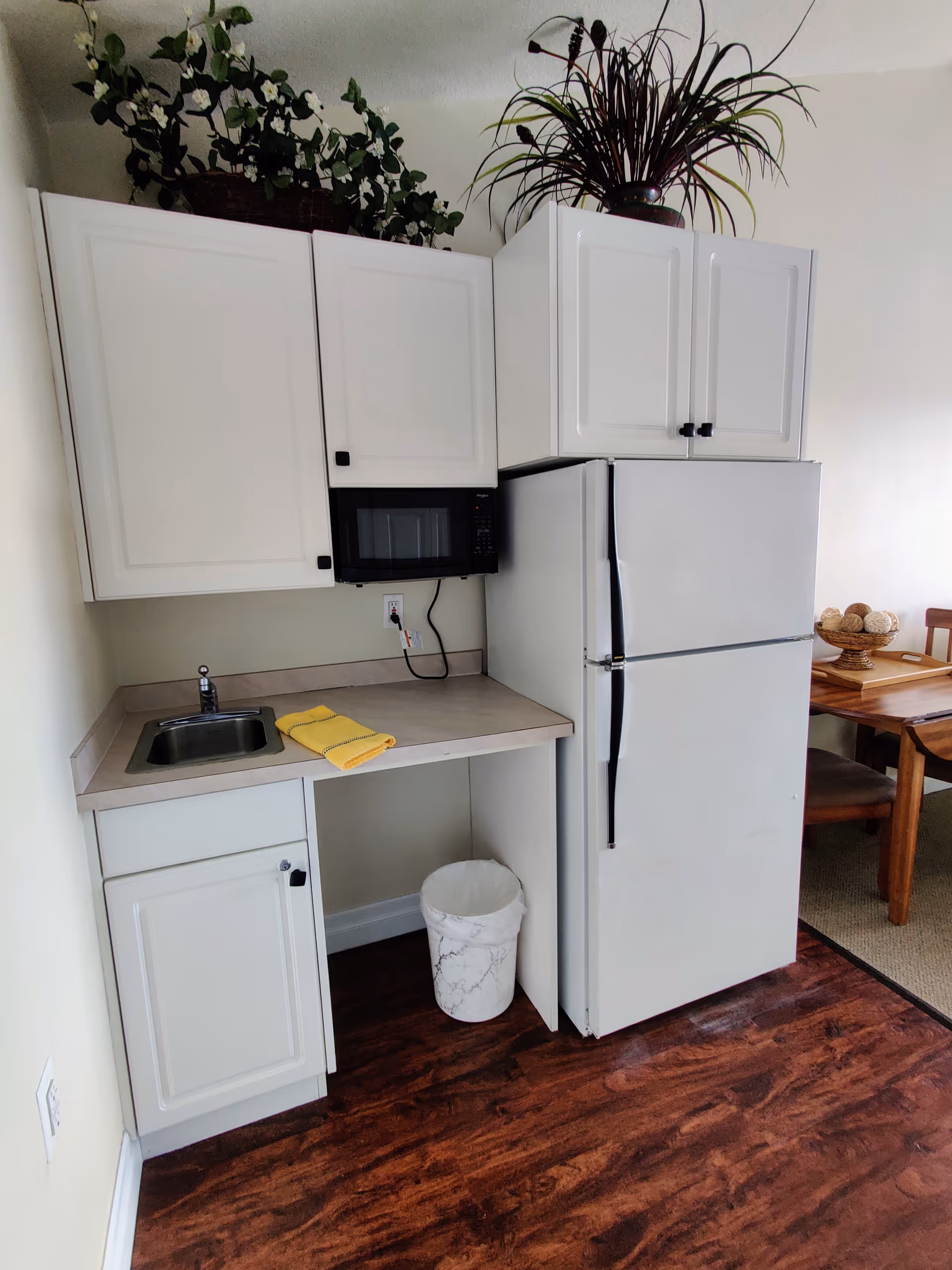 Small kitchen area with white cabinets, a countertop with a sink, a microwave mounted under the upper cabinets, a white refrigerator, and a trash can underneath the counter. There are two potted plants on top of the cabinets and a yellow towel on the counter. Part of a wooden dining table with chairs is visible to the right.