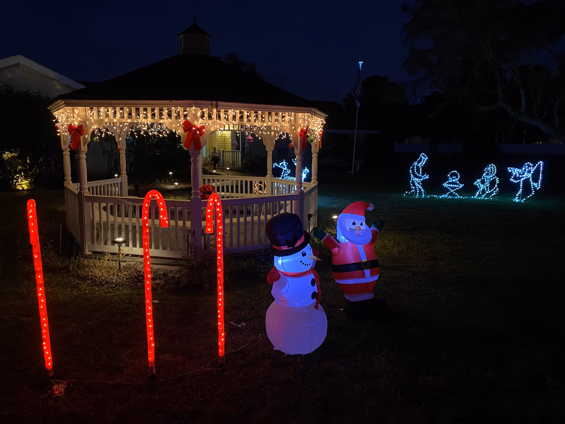 A gazebo decorated with white string lights and red bows at night. In front of the gazebo are three red candy cane-shaped lights and inflatable holiday figures of a snowman and Santa Claus. In the background, blue lights form a nativity scene with figures of people and an angel on a grassy lawn.