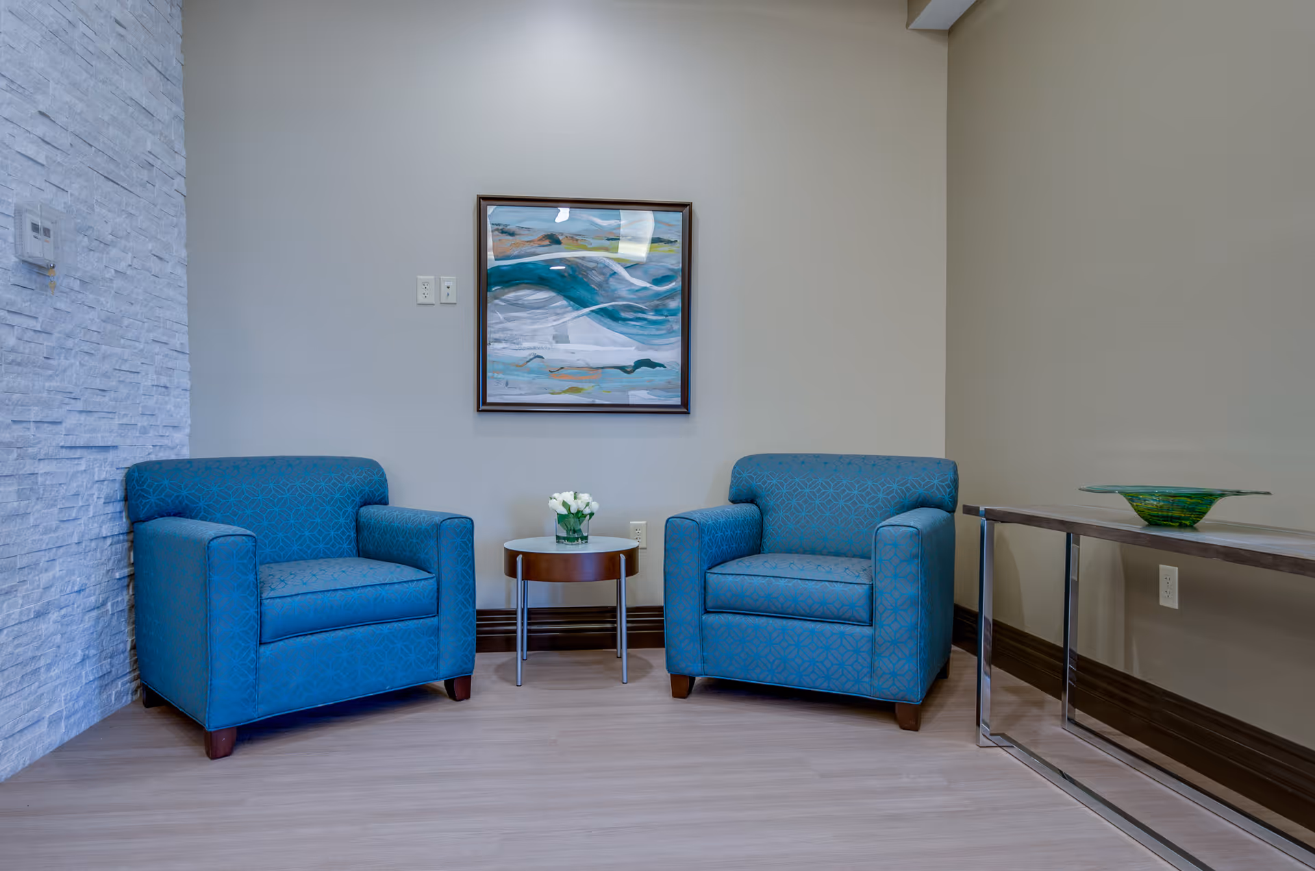 A cozy seating area with two blue upholstered armchairs separated by a small round table holding a vase of white flowers. Above the table hangs a framed abstract painting with blue and green tones. To the right, there is a narrow console table with a decorative green glass bowl. The walls are painted beige and one wall has a textured white stone finish.