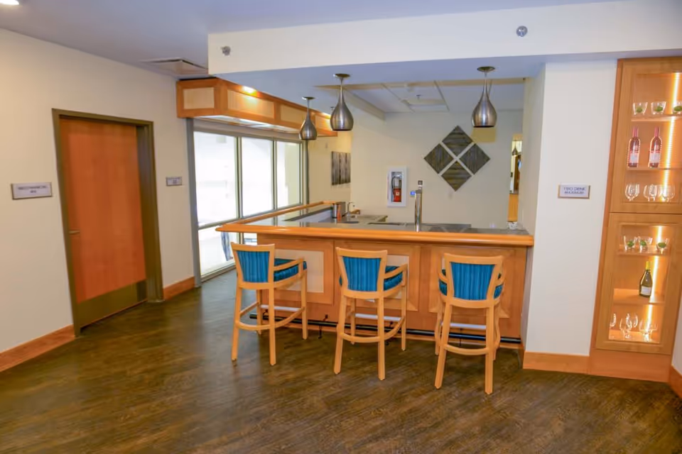 Small indoor bar area with three wooden barstools at a counter, pendant lights, and a glass-front display cabinet.