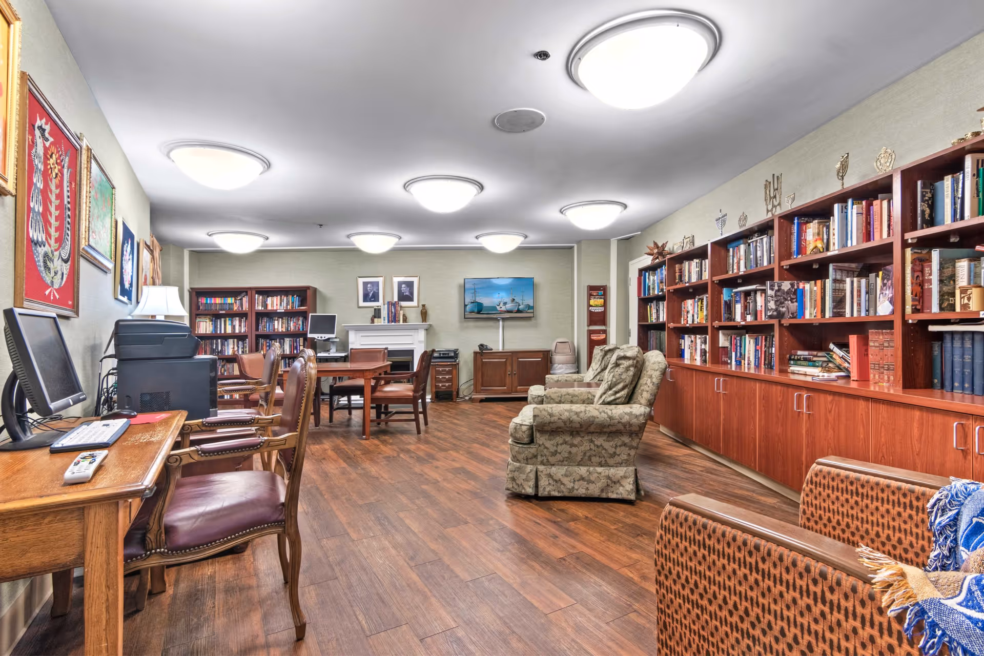 A cozy and well-lit common room with wooden flooring, bookshelves filled with books, comfortable armchairs, a table with chairs, a computer workstation, and framed artwork on the walls. The room has ceiling lights and a television mounted on the far wall.