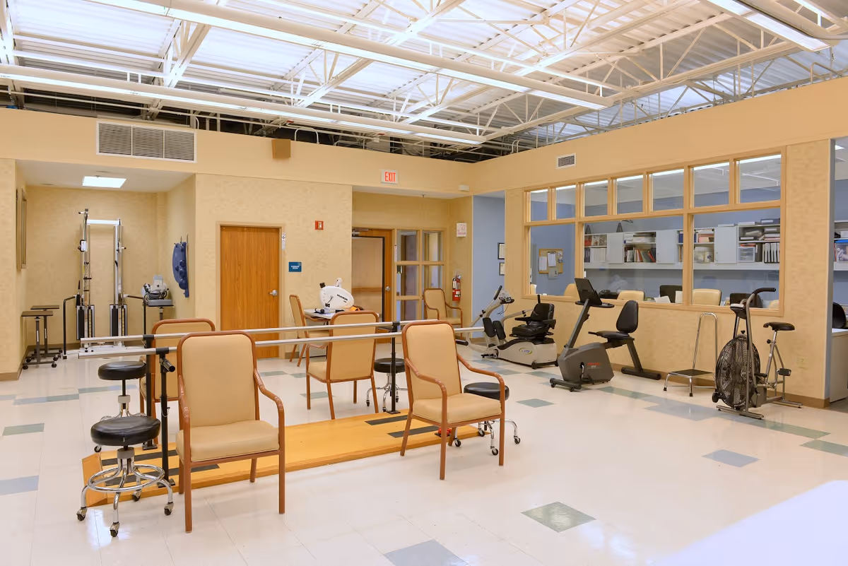 A rehabilitation room with exercise equipment including stationary bikes, parallel bars for walking practice, chairs, and stools. The room has a high ceiling with exposed beams and a large window looking into an adjacent office or workspace with shelves and cabinets.