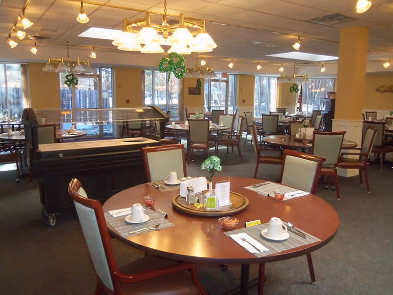 A spacious dining room in an assisted living facility with multiple round tables set for meals. Each table has placemats, cups, utensils, and small centerpieces. Large windows allow natural light to fill the room, and there are overhead light fixtures. Some green shamrock decorations are hanging from the ceiling, suggesting a festive atmosphere.