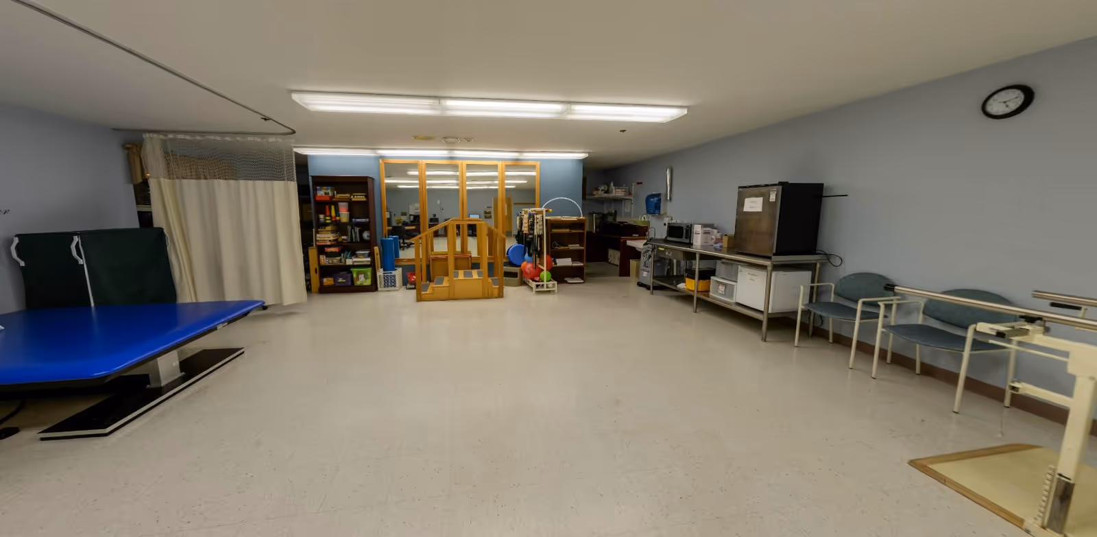 A spacious room with light blue walls and white tiled floor, featuring a blue therapy table on the left, a wooden ramp and exercise equipment in the center, shelves with supplies, a metal table with a microwave and refrigerator on the right, and several chairs along the right wall. A clock is mounted on the wall above the chairs.