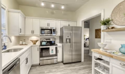 Modern kitchen with white cabinets, stainless steel refrigerator, oven, microwave, and sink. Light wood flooring and a small counter area with decorative items and plants. A doorway leads to a bedroom in the background.