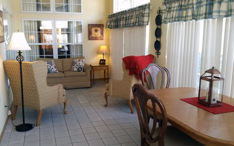 A cozy interior room with a tiled floor featuring a beige sofa with patterned pillows, two beige armchairs, a wooden dining table with chairs, a floor lamp, a small side table with a lamp, and vertical blinds covering large windows.