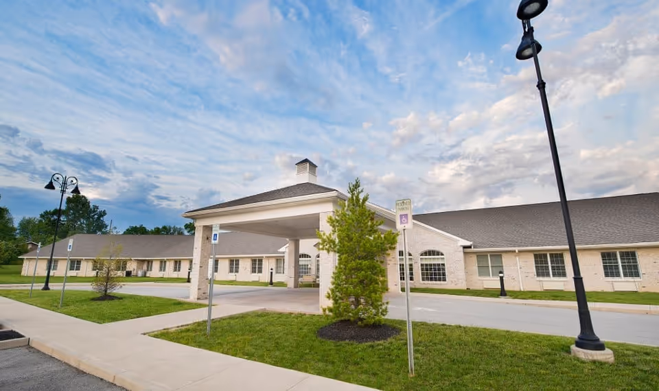 Front exterior of a single-story brick senior living facility with a covered drive-through entrance, lamp posts, and a small landscaped lawn.